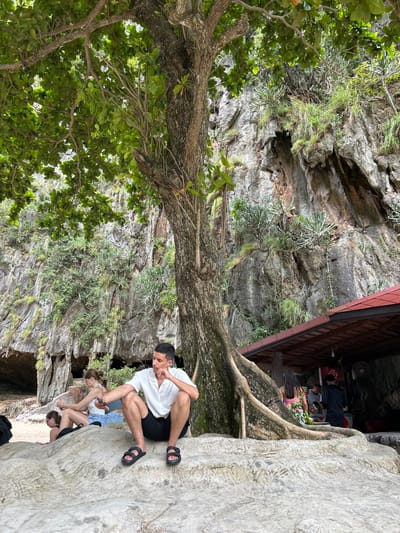 In Krasom, Thailand, a group of people are gathered under the shade of a large tree on a rocky outcrop near a cliff face. The man in the foreground, wearing a white short-sleeved shirt and black shorts, sits on a large, light-colored rock, looking pensive with his hand on his chin. Behind him, a woman with blonde hair tied back, wearing a white top and blue shorts, sits on another rock, looking at her phone. To her left, another woman with dark hair, wearing glasses and a black top, is also seated on the rocks. In the background, a reddish-brown roofed structure, possibly a food stall or shop, is visible against the imposing cliff face which is covered in lush green vegetation and shows openings that suggest caves or overhangs. The roots of the large tree are prominently visible, spreading across the rocks. Several other people are seen near the structure in the background, and one person is standing near the tree on the right side of the frame, partially obscured. The environment appears to be a natural, tropical setting, likely a beach or a scenic viewpoint, with the ocean possibly out of frame.