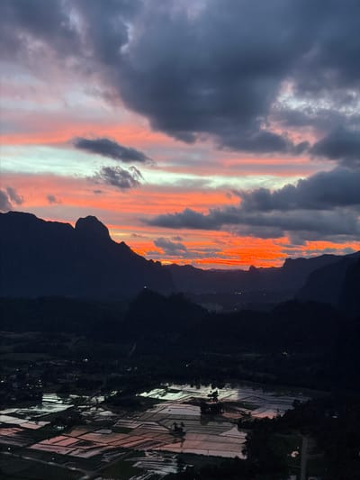 The image captures a dramatic sunset over the karst landscape of Vang Vieng, Laos. The sky is a vibrant spectacle of oranges, pinks, and grays, with dark, brooding clouds dominating the upper portion of the frame. Below the tumultuous sky, silhouetted mountain peaks rise majestically, their jagged outlines a stark contrast against the colorful horizon. In the foreground, a patchwork of flooded rice paddies reflects the fading light, creating a mosaic of muted colors and shimmering surfaces. The landscape appears rural and tranquil, with minimal signs of human presence, save for distant lights suggesting scattered dwellings. The overall impression is one of natural beauty and serene, untouched wilderness.