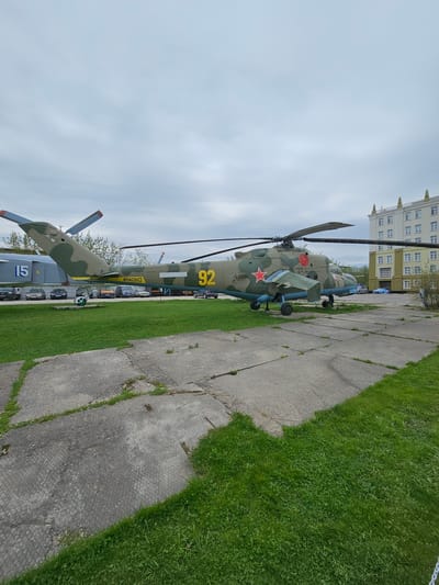 The image captures a military helicopter, prominently displayed on a cracked concrete surface adjacent to a grassy area. The helicopter is painted in a camouflage pattern with a red star insignia and the number "92" in yellow. In the background, there is a multi-story building under a cloudy sky. Based on the location, this photograph was likely taken at the Vadim Zadorozhny Museum of Technology in Arkhangel’skoye, Russia.