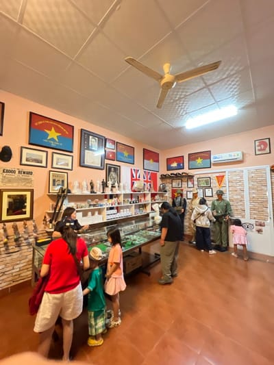 A group of people are looking at a display case in a room decorated with various flags and memorabilia. The room has a light peach-colored wall adorned with framed pictures, flags, and posters. A ceiling fan hangs in the upper left corner. The display case appears to contain historical artifacts or collectibles. The floor is a reddish-brown hue.