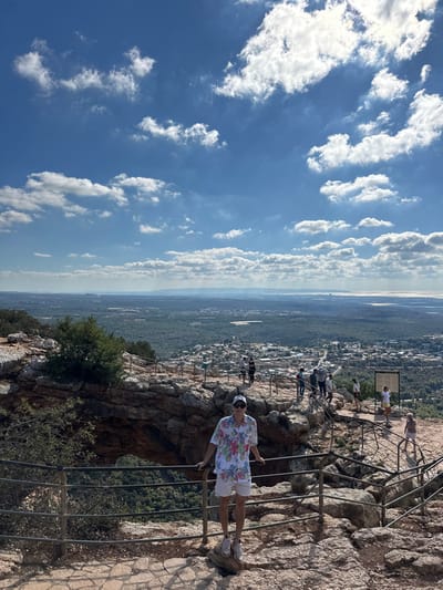The image presents an elevated panoramic view from a rocky lookout point within the Maale Yosef Regional Council, Israel, under a bright blue sky with scattered white cumulus clouds.
In the foreground, a male subject stands centrally, facing the camera. He is wearing a white short-sleeved shirt with a multicolored floral pattern, light-colored shorts, white athletic shoes, a white baseball cap, and sunglasses. He stands on an uneven rocky surface, gripping a dark metal guardrail that delineates the edge of the cliff. Behind him, a natural rock arch formation is visible, framing a deep, forested valley below.
The mid-ground features a rugged, rocky terrain with established pathways and additional sections of dark metal guardrails. Approximately eight to ten other individuals are present on these paths, scattered across the viewpoint. They appear to be tourists or hikers, standing or walking, observing the distant scenery. An informational sign on a metal stand is positioned on the right side of the path. Sparse green vegetation, including small trees and bushes, grows among the rocks.
The background displays an expansive landscape. Immediately below the lookout, a dense forested valley transitions into a residential area with numerous buildings and a road network, indicative of a settlement. Beyond this, vast stretches of green forests extend towards the horizon, where distant hills or mountains are visible. To the far right, a body of water, consistent with the Mediterranean Sea, meets the horizon line. The scene is illuminated by strong, natural daylight.