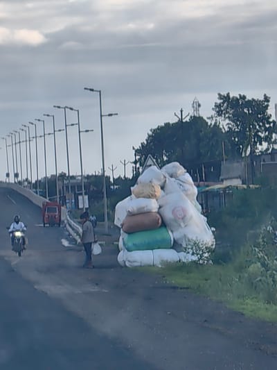 The image depicts a street scene in Malkapur, India. A long row of streetlights stretches into the distance along the left side of the road, which curves upwards. In the foreground, a large pile of white and green sacks is stacked on the right side of the road. A man in a grey shirt and dark pants, holding a white plastic bag, walks towards the left of the frame. A person on a scooter, wearing a white shirt and black pants, is riding on the left side of the road, heading away from the viewer. Further down the road, a small orange vehicle is visible. In the background, trees and buildings are scattered, with a cell tower visible above the trees. The sky is overcast with grey clouds. The overall impression is that of a typical roadside scene in an Indian town, with elements of transportation, commerce, and infrastructure.