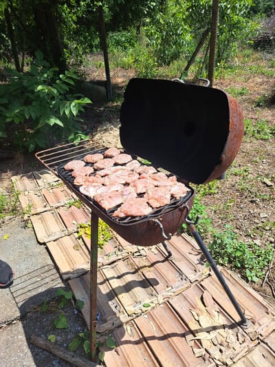 The image shows a rusty barrel grill with many pieces of raw meat on its grates, cooking outdoors in what appears to be a backyard or garden area. The grill is placed on an uneven surface covered with old terracotta roof tiles. In the background, there are trees and vegetation, with a wire fence visible in the distance. The ground is covered with dirt, grass, and some debris. A shadow from the grill and potentially a person is cast on the tiled surface. There is a black shoe with a red sole visible in the bottom left corner of the image. The scene suggests an informal outdoor cooking setting, possibly during a summer day.