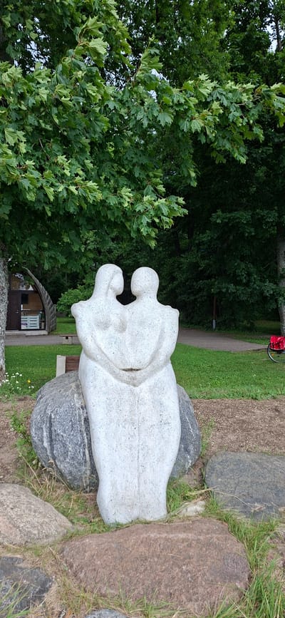 In Valgeranna, Estonia, a modern abstract sculpture of two figures embracing is positioned on a large, grey boulder. The sculpture, rendered in a light-colored stone, depicts the figures with simplified, flowing forms, their heads tilted towards each other as if in a tender moment. They are situated in an outdoor setting with a backdrop of lush green trees and a manicured lawn. To the left of the sculpture, a small, curved wooden structure with a window and door is visible. A wooden bench is placed behind the sculpture. In the background, a paved pathway winds through the grassy area, and a red bag is placed on the back of a bicycle. The foreground is characterized by rough, brown and grey rocks with scattered grass.