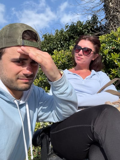 Two individuals, a male and a female, are seated on a black metal park bench in an outdoor setting within Kenilworth, United Kingdom. The male, positioned on the left and closer to the viewer, is young, wearing an olive green mesh-back baseball cap, a light blue hooded sweatshirt, and a gold-colored watch on his left wrist. He holds his right hand up, shading his eyes from direct sunlight, and looks forward. The female, seated to his right and slightly behind him, is wearing dark sunglasses, a light blue ribbed long-sleeve top, and black leggings. Her arms are crossed, and she looks towards her right. A light brown shoulder bag rests beside her. The background consists of dense green foliage and some bare tree branches under a clear blue sky with scattered white clouds. Strong natural light illuminates the scene, creating distinct shadows.