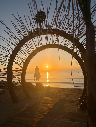 The image shows a sunset scene on a beach in Trikomo, Northern Cyprus. In the foreground, there is a wooden walkway leading to the beach, with sand on either side. A large, circular metal archway with radiating metal rods is positioned centrally. The word "PERA" is visible in letters across the top of the archway. Inside the archway, a beach umbrella is folded and leaning on a lounge chair. The sun is setting over the calm sea, casting a golden reflection on the water. The sky is clear with a gradient of blue and orange hues. A palm tree is visible on the right side of the frame, with its fronds extending towards the archway.