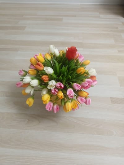 A bouquet of artificial tulips sits on a light-colored wooden floor. The tulips are a mix of colors, including pink, yellow, white, orange, and one red tulip stands out at the top of the arrangement. The bouquet is lush and full, with green leaves interspersed among the flowers. The wooden floor has a visible grain pattern and appears to be made of planks. The lighting suggests an indoor setting, possibly a home or an office, with no indication of time of day or weather. There are no people or animals visible in the image, and no text is present. The overall mood is simple and decorative, highlighting the colorful floral arrangement against a neutral background.