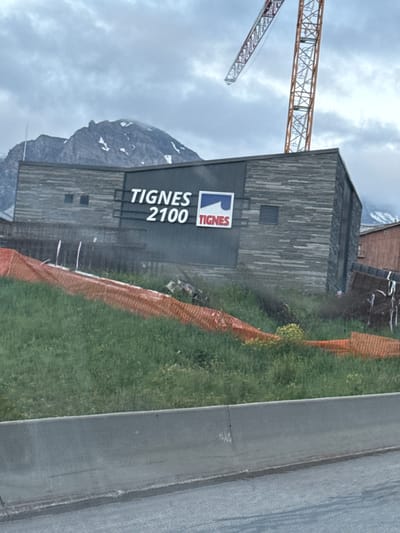In Tignes, France, a building with a dark, horizontally paneled facade displays signage for "TIGNES 2100" with a blue and red logo featuring a stylized wave and the word "TIGNES". The building is situated in front of a mountainous backdrop with snow-capped peaks under a cloudy sky. A large yellow and red construction crane is visible behind and to the right of the building. Orange safety netting is erected in front of the building, bordering a grassy, overgrown area. A concrete barrier runs along the bottom foreground of the image.