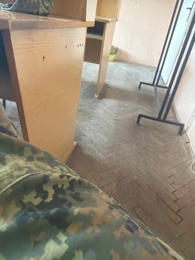 The image shows an indoor scene with a partial view of a person wearing camouflage clothing in the foreground. The background includes a table or desk with a metal frame and a white surface. A wooden structure resembling a cabinet or desk is partially visible on the left side of the frame. The floor is tiled in a herringbone pattern. The location is unknown and no specific details suggest the scene is in Dolna Mitropolia, Bulgaria.