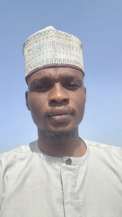 The scene is depicted in Badiko, Nigeria. A dark-skinned adult male is positioned centrally, facing directly towards the viewer. He possesses a short, dark beard and stubble on his chin and jawline. He wears a tall, light-colored embroidered cap with intricate geometric patterns, featuring a dark reddish-brown band at its base. His clothing consists of a light grey or beige long-sleeved garment with a round collar and a partial button placket, secured by a visible dark button at the collar. The background consists solely of a clear, light blue sky, indicating an outdoor setting with no discernible additional features.