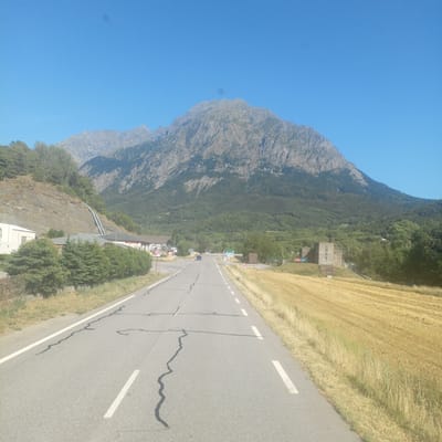 The image shows a scenic view of a road leading towards a large, rocky mountain under a clear blue sky. The road, asphalt, has white dashed lines on the right side and solid white lines on the left side, with some cracks and patching visible on the surface. To the left of the road, there is a steep, rocky embankment with some trees and a building with pipes running up the side of the embankment. Further down the road, there are some low-lying buildings, including one with a sign that reads "ZONA TONS". A dark-colored car is driving away from the camera on the road. To the right of the road, a field of dry, golden grass or crops stretches towards the background. In the middle ground, there is a rectangular concrete building on a small hill surrounded by trees. The mountain in the background is covered in vegetation at its base, transitioning to bare rock towards the summit. The overall scene depicts a rural or mountainous area, likely in the vicinity of Saint-Firmin, France, given the context provided. There are no people clearly visible in the image. The lighting suggests it is daytime, likely mid-morning or afternoon, with the sun casting shadows.