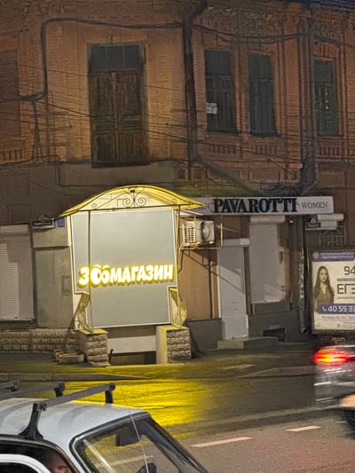 This image depicts a nighttime scene on a street in Chemul'ga, Russia. The perspective is from a vehicle looking towards a brick building with several windows. The building's facade is aged, with visible wires and some signs.
Prominently featured is a glowing yellow sign with Cyrillic text that reads "ЗООМАГАЗИН" (ZOO-MAGAZIN), indicating a pet store. Next to it, a vertical sign displays "PAVAROTTI WOMEN" in white letters on a dark background, suggesting a women's clothing store. Above these, other signs are partially visible, including one with the text "РЕСПУБЛИКАНСКИЙ ЦЕНТР ПОДГОТОВКИ К ЕГЭ" (Republican Center for Exam Preparation) and a phone number "40 55 33". A street sign with "33 Маяковского" (33 Mayakovsky) is also present.
The street itself is wet, reflecting the bright yellow and red lights from the signs and possibly from passing vehicles. The presence of a car in the foreground, with its door open, suggests someone is either exiting or entering it, or perhaps interacting with the store. The overall atmosphere is that of a typical urban street at night, with artificial lighting illuminating the storefronts and casting reflections on the wet pavement. The time of day is clearly night due to the darkness and the illuminated signs.