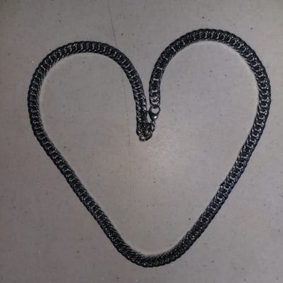A silver-colored metallic chain is displayed on a light-colored, speckled surface. The chain is arranged into a distinct heart shape, with its two ends meeting at the top center. A lobster clasp and an extender chain are visible at the junction of the chain ends. The chain itself features interlocking, flattened links, consistent with a curb chain design. The underlying surface is uniformly light-toned, appearing off-white or light gray, and exhibits a consistent distribution of fine, reflective particulate matter across its plane. The scene is illuminated by an overhead light source, creating subtle reflections and localized shadows on the chain. No individuals or other primary objects are present within the frame. This image is documented to represent a scene in Gimpo-si, South Korea.