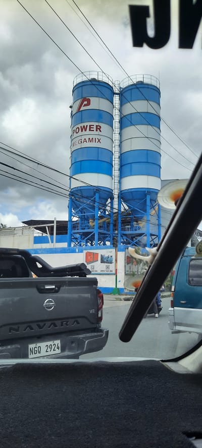 The image captures a view from inside a vehicle, looking forward towards two large industrial silos. These silos are painted with alternating blue and white bands and have a large "P" logo with the word "POWER" and "MEGMIX" printed on them. The silos are supported by a blue metal structure. In the foreground, the rear of a dark gray Nissan Navara pickup truck is visible, with its license plate reading "NGQ 2924". To the right of the Navara, part of a blue van is visible. In the background, beyond the silos, there are buildings and infrastructure, suggesting an industrial area. The sky is overcast with clouds. A few electrical wires are stretched across the upper portion of the image. The scene appears to be on a road, with other vehicles present and the general atmosphere of a moderately busy urban or industrial environment. The location is identified as Kawit, Philippines, implying a specific geographical context for this industrial setting.