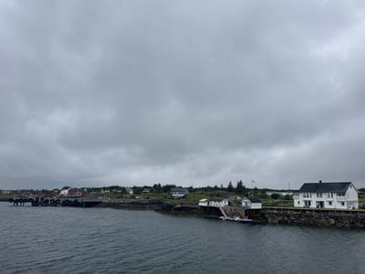 This is a medium-distance, eye-level, outdoor shot of a coastal town in Sleneset, Norway, under a cloudy sky. The foreground is dominated by dark, choppy water, with the camera positioned at or near water level. In the midground, a rocky shoreline with some vegetation is visible, leading to a settlement of buildings.
On the left, a pier extends into the water, lined with large black tires for protection. Several buildings, mostly red and white with dark roofs, are situated along the pier and the shoreline. Some of these appear to be houses, while others might be storage or industrial structures. Cars are parked on the land behind the pier.
Further to the right, a prominent white two-story house with dark windows and balconies stands on a rocky outcrop overlooking the water. A smaller white building, possibly a boathouse or annex, is attached to the main house and sits on a wooden pier extending over the water. A small blue boat is docked at this pier. Trees are scattered throughout the landscape, with more dense greenery visible in the background. A flagpole with a flag can be seen flying near the white house. The overall scene is one of a quiet, coastal community.