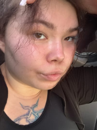 The media file is a close-up, horizontal, slightly tilted photograph of a person's face and upper chest. The subject appears to be a young woman with brown hair that is falling across her face, obscuring part of her forehead and eye. She has brown eyes, a light complexion, and her lips are slightly parted. A tattoo in shades of blue and purple, resembling a lightning bolt or abstract pattern, is visible on her upper chest. She is wearing a dark, ribbed garment that appears to be a top or sweater. The background is dark and out of focus, suggesting an indoor setting, possibly with some reflective surfaces. The lighting is soft, illuminating her face. There is no visible text in the image, and no clear indicators of the time of day or weather. The overall impression is a candid, personal snapshot.