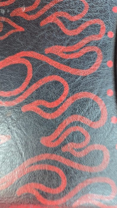 A close-up image shows a black surface adorned with a pattern of stylized red flames. The flames are depicted as rounded, elongated shapes that twist and curve across the surface. Small red dots are visible near the upper edge. The black base appears to have a textured, possibly leather-like, quality. The red shapes are applied with a slightly rough edge, suggesting paint or a similar medium.