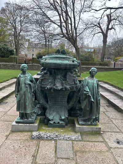 Here's a description of the image:
The image shows the William Kelly Memorial in Aberdeen, United Kingdom. The monument is made of bronze and features two statues of women standing on either side of a large central structure covered in intricate carvings and text. The statues and structure are mounted on a stone base.
The monument sits on a paved area with grass and trees surrounding it. In the background, there are buildings and foliage. The sky appears overcast.