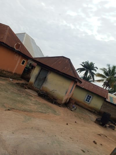 The image depicts a cluster of residential buildings with corrugated iron roofs in Gindin Dutse, Nigeria. The structures vary in color, with one prominent building painted orange and another in shades of yellow and beige. A rusted satellite dish is visible on the orange building's roof. The ground is a mixture of dirt and sand, with some scattered debris. In the foreground, a small chicken is walking across the dirt path. Palm trees are visible in the background, suggesting a tropical or subtropical environment. The sky is overcast with clouds. There are two large metal barrels and several rocks near one of the buildings, one barrel appearing to be a water drum and the other a stove. A small, square window with white panes is set into the yellow building. A metal door with vertical slats is present on the yellow structure.