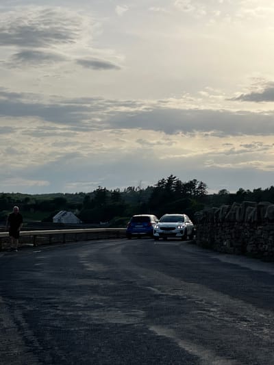 The image depicts a road alongside a stone wall and a wooded area, under a cloudy sky. Two cars are parked on the side of the road: a white SUV and a blue hatchback. In the foreground, a person stands near the edge of the road. The scene appears to be set in Terhillion, Ireland, given the provided location context. The lighting suggests it is either early morning or late afternoon, with the sky filled with diffuse light and textured clouds, hinting at overcast or possibly rainy weather. The stone wall, the rolling hills in the background covered with trees, and the presence of a small white building suggest a rural or semi-rural setting. The overall mood is calm and perhaps a bit somber due to the overcast sky. No visible text is present in the image.
