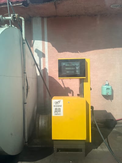 This is a top-down, slightly angled outdoor photograph taken in Yelyzavetivka, Ukraine. The image captures a yellow fuel dispenser with a digital display and a large, white cylindrical tank. The fuel dispenser has text in Ukrainian, including "ВАРТІСТЬ" (cost), "ОБ'ЄМ" (volume), and "ЦІНА" (price), with displayed values of 999.60 UAH, 14.70 liters, and 68.00 UAH/liter respectively. A logo with the text "KCEM ДП euro5" is also visible on the dispenser.

In the background, a light green plastic container with a white tap is situated on a textured, pinkish-brown wall. A significant shadow cast by an unseen object falls across the scene from the right, suggesting it's daytime with bright sunlight. The ground appears to be paved with a reddish material and has some white stripe markings. The overall setting suggests a fuel station or an industrial area. There are no people visible in the frame, and the mood of the image is functional and utilitarian. The focus is on the fuel dispensing equipment and the storage tank.