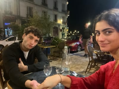 This nighttime image captures an outdoor dining scene in the city of Orléans, France.
In the immediate foreground, two young adults are seated at a dark-topped outdoor table. On the left, a male with dark, curly hair and a neutral expression looks towards the camera. He wears a black zippered long-sleeved top over a white shirt. His right hand is holding the left hand of the female seated opposite him. On the right, a female with dark hair smiles faintly towards the camera. She is wearing a red knitted top and a beaded necklace.
On the table between them, there are two empty stemmed wine glasses, one clear water glass, and a clear glass bottle containing water. Cutlery is visible next to the male's position.
In the mid-ground, behind the seated individuals, a male adult in a dark short-sleeved shirt and patterned shorts stands on a paved surface. A red four-door hatchback vehicle, likely a Peugeot 208, is parked directly behind him. Another white car is partially visible on the far left.
The background consists of multi-story buildings with light-colored facades and closed dark shutters, typical of European architecture. A tree with green foliage is situated between the buildings and the street level. The scene is illuminated by streetlights and ambient light from establishments, casting soft shadows. The overall environment suggests a lively street or public square at night.