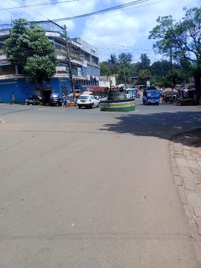 The image depicts a sunlit urban intersection in Murang'a, Kenya, under a blue sky with scattered white clouds. The foreground shows an asphalt road surface, transitioning to a segmented concrete sidewalk on the far right. Centrally, a circular concrete traffic island is painted with horizontal green, yellow, and black stripes; it features a tiered, dark grey structure on its top.

On the left side of the intersection, a multi-story building with a blue lower facade is visible, displaying a "BOOK CENTRE" sign. A large, dense-leafed green tree stands directly in front of this building. Multiple utility poles, heavily laden with numerous overhead wires, rise along the left side of the road and extend across the frame.

Vehicle traffic includes a white sedan with a visible rear license plate, positioned near the traffic island and facing away from the viewer. A dark blue sedan is parked closer to the blue building. In the right background, a blue light-duty truck approaches the intersection. Additional indistinct vehicles and several market stalls with colorful umbrellas are visible further in the background to the right.

Pedestrian presence includes one individual in a light-colored garment standing on the far-left sidewalk, and multiple other individuals near the blue building and the dark blue sedan. The scene is brightly illuminated, casting distinct shadows on the asphalt from surrounding structures and trees.