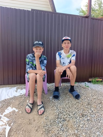 Two young boys are sitting on pink plastic chairs in front of a brown corrugated metal fence. The boy on the left is wearing a black baseball cap with white text that reads "OFFLINE," a dark t-shirt with a black, white, and green graphic print, and dark shorts. He is sitting with his legs crossed and his left hand is covering his mouth. The boy on the right is wearing a blue and white baseball cap, a white t-shirt with a blue and green graphic print, and dark shorts. He is sitting with his legs apart, his hands resting on his knees. Both boys are wearing dark socks and sandals.

The ground in front of them is covered with gravel and some sand. To the left of the boys, there is some white material, possibly landscaping fabric, partially covered with sand and debris. The fence behind them is tall and appears to be a boundary for a yard or property. The sky is visible above the fence, with some clouds and a hint of trees. The lighting suggests it is daytime, likely late morning or afternoon, with bright sunlight casting shadows. The overall scene appears to be an outdoor residential setting, possibly in a rural or suburban area. The boys have neutral to slightly amused expressions.