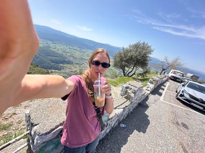 A woman is holding a Starbucks drink in front of a valley landscape. She wears sunglasses and a pink shirt. A car is parked nearby on a paved surface. The scene is bright and sunny with blue sky and scattered clouds. The location is in the city of Ula, Turkey.
