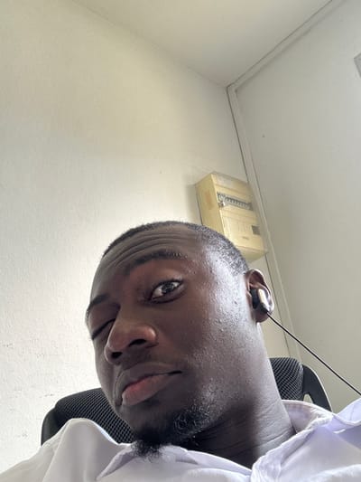 This low-angle, close-up photograph captures a man, likely in a selfie, from a perspective looking upwards. The man, with dark skin and short dark hair, is positioned diagonally in the frame, with his head tilted. He has a subtle smile or smirk on his face and looks directly into the camera with his right eye.

He is wearing a white collared shirt, visible around his right shoulder and chest. A black mesh office chair is partially visible behind his shoulder, suggesting he might be seated. An earphone or headset with a thin black wire is inserted into his right ear.

The background features light-colored, possibly off-white or beige, walls and a white ceiling. In the upper left portion of the background, mounted high on the wall, is a light-yellowish electrical or utility box. It appears somewhat aged or dusty, with internal components resembling switches or gauges visible through its translucent cover. The setting is an indoor space, possibly an office or a functional room. The ambient lighting suggests it is daytime. Based on the provided context, this scene is in Abidjan, Ivory Coast. There is no visible text in the image.