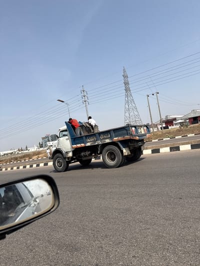 The image captures a bright daytime scene from a vehicle on a road in Aso Housing Estate, Nigeria. The primary subject is a light blue dump truck traveling in the same direction, positioned on the right side of an asphalt road. Two men are visible in the truck's open bed, riding amidst cargo; one wears a white shirt, the other an orange shirt.
The road features distinct black and white striped curbs along its right edge. Overhead, a clear, light blue sky is traversed by numerous power lines. A large metal lattice transmission tower (pylon) stands prominently on the left side of the frame, accompanied by smaller utility poles. In the background, various buildings and some sparse vegetation line the roadside. The lower right corner of the image shows the side mirror of the vehicle from which the photo was taken, reflecting parts of the vehicle's interior and the road behind.
Visible text includes "GOODS" painted on the side of the blue truck bed, and "OZZO" on the rear fender. Another word, possibly "GANIYO," is also present on the truck. The bright, clear sky suggests sunny weather conditions.