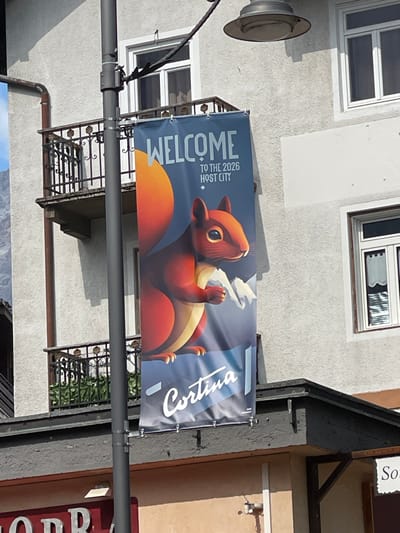 The image shows a banner hanging on the side of a building, likely in Cortina d'Ampezzo, Italy. The banner features a cartoonish red squirrel with large eyes, looking upwards and holding something white, possibly smoke or steam, in its paws. The banner has the text "WELCOME TO THE 2026 HOST CITY" in white and blue lettering on the left side, and "Cortina" written in a stylized script on the right side. The building itself is light-colored with windows and a balcony with black metal railings. A street lamp is visible on the left, and a dark pipe runs along the building's facade. The scene appears to be outdoors during the daytime, with sunlight casting shadows, suggesting a clear day. The overall impression is one of anticipation and welcome for the upcoming 2026 event in Cortina.