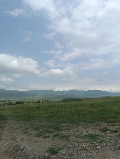 The image depicts a rural landscape with rolling green hills and mountains in the background under a partly cloudy sky. In the foreground, a dirt path with gravel is visible, leading to a grassy field dotted with wildflowers. The horizon is marked by a range of low, verdant mountains. The scene appears to be a serene, natural environment, likely in a rural area such as the village of Hartagyugh, Armenia, given the geological and vegetation characteristics. There are no people or animals visible in the image.