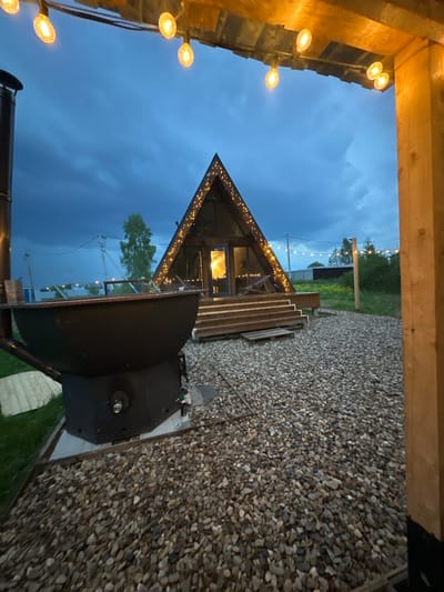 The image shows a view from a wooden structure, possibly a porch or entryway, looking towards a small A-frame cabin at twilight. The cabin is decorated with string lights along its roofline. The surrounding ground is covered with gravel. A large, dark metal object, potentially a grill or smoker, is situated on a concrete slab next to the gravel area. In the distance, there are other buildings and trees. String lights are visible in the background. The location is Малеевка, Russia.