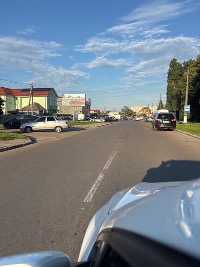 The image shows a street scene in Pereiaslav, Ukraine. The perspective is from a car looking forward along the road. Several cars are parked along the right side of the road, and traffic is visible further down. Buildings line the street, some with red roofs and green walls, and solar panels are visible on one roof. A large billboard is mounted on a building on the left side of the street.
The sky is blue with scattered white clouds. The sun is bright, indicating it is daytime, likely late afternoon due to the long shadows. The weather appears to be clear and sunny. A bus stop sign is visible on a pole at the beginning of the street. There are power lines running along the street.
The overall impression is of a quiet residential or commercial street on a pleasant day. There are no people clearly visible in the image. The scene is static, with cars parked and the general atmosphere of everyday life.
Visible text:
- "+3 8063 820 41 27" and "Впевнені для блиску вікон" (likely an advertisement for window cleaning services) on the billboard.
- "АПТЕКА" (Pharmacy) is visible on a building.