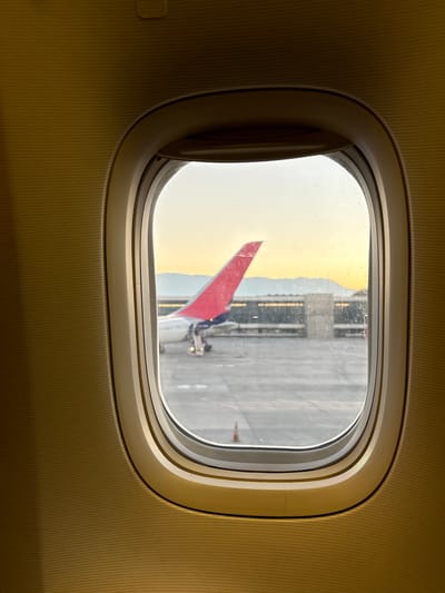 The image shows a view through an airplane window. A red and blue tail fin is visible, likely belonging to another plane on the tarmac. The outside environment appears to be an airport, possibly with a building in the background. It is day time. Based on the shape of the fin, this is a Delta Airlines aircraft. The image does not contain information to verify it is located in Pudahuel, Chile.