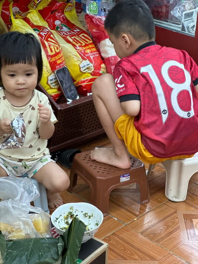 In Bắc Yên, Vietnam, two children are seated on plastic stools. The younger child, a girl with dark hair and bangs, wears a light-colored t-shirt with heart patterns and patterned shorts. She holds a silver foil wrapper. The older child, a boy, is dressed in a red jersey with a large "18" printed on the back and yellow shorts. He is facing away from the camera. They are surrounded by large yellow and red bags, likely containing rice, with Vietnamese text. A smartphone lies near the younger child. Food items, including a bowl with remnants of a dish and a banana wrapped in banana leaves, are visible on the floor and a box. The environment appears to be a market or store, with tiled flooring and shelves in the background.