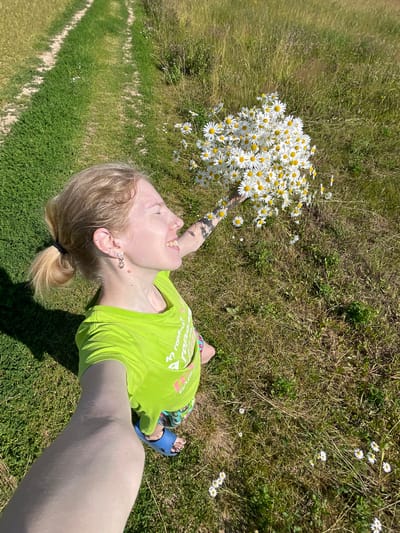 A young person with blonde hair, wearing a bright green t-shirt and blue sandals, lies on their back in a grassy field, holding a large bouquet of daisies. The person is looking upwards with their mouth open in what appears to be laughter or a shout of joy. Their hair is pulled back into a ponytail, and they have several piercings in their ears and a tattoo on their arm. The bouquet of daisies is voluminous and takes up a significant portion of the frame.
The setting is a sunny outdoor environment with a mix of green grass and drier, brown vegetation. Wild daisies are scattered throughout the field. In the background, a pathway or mowed strip of grass runs diagonally through the scene, suggesting a park or a rural area. The lighting indicates it is daytime, likely a sunny afternoon, given the shadows. The overall mood is cheerful and carefree, with the person enjoying the flowers and the natural surroundings. The text on the t-shirt is partially visible and appears to be in Cyrillic script. The location context is Сям-Можга, Russia.