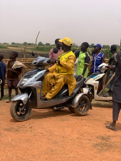The scene is a dirt road in Ringimawa, Nigeria. A man wearing a yellow vest with the word "WOFAN" on it is sitting on a silver three-wheeled motorcycle. There is another WOFAN motorcycle behind him. Several other people are standing around the motorcycles, including a young boy with a white shirt and a young man wearing a yellow shirt and a black vest. Behind the group, there is a green field with a fence. The man on the motorcycle is facing forward. The sun is out and there is a clear sky. The ground is dirt with some weeds. There is a stick lying on the ground to the left of the motorcycles. The man on the motorcycle is looking straight ahead. The other people are looking in various directions.