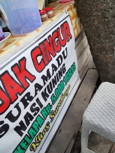 The image captures a close-up view of a white banner with red, black, and green lettering. The banner appears to be hanging outdoors, possibly at a food stall, given the presence of a plastic water dispenser, small jars with lids, and a textured plastic stool in the background. A large tree trunk is also visible to the right, with its rough bark taking up a significant portion of the frame. The banner itself advertises a product called "JUAL CINGUR MADU NASI KUNING Rp. 5.000 1GELAS KHAS SURABAYA," which translates to "For Sale: Cingur, Honey, Yellow Rice Rp. 5,000 1 Glass, Specialty of Surabaya." The lettering is bold and eye-catching. The background suggests a somewhat rustic or informal setting. The overall lighting indicates it's daytime. The provided crops highlight specific sections of the banner and the surrounding objects. While the image is taken in Palangka Raya, Indonesia, no specific landmarks or contextual elements within the scene confirm this location.