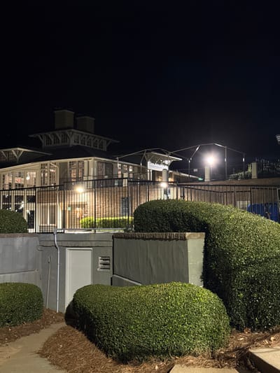 The image shows a multi-story building at night, illuminated by streetlights. The building has white balconies and railings. A tall fence runs along the front, partially obscured by neatly trimmed, rounded bushes. The sky is dark, and the ground is covered in pine needles and pavement. The scene is urban, combining architectural and landscape elements.
