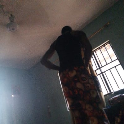 A man stands with hands on his hips, facing away from the viewer, inside a room. He wears a dark t-shirt and a long skirt with a floral pattern. A window with metal bars is to his right, allowing daylight to stream in. A ceiling fan is visible in the upper left corner. The room is dimly lit, with a light-colored ceiling and greenish walls. The focus is on the man's silhouette and the patterned skirt.