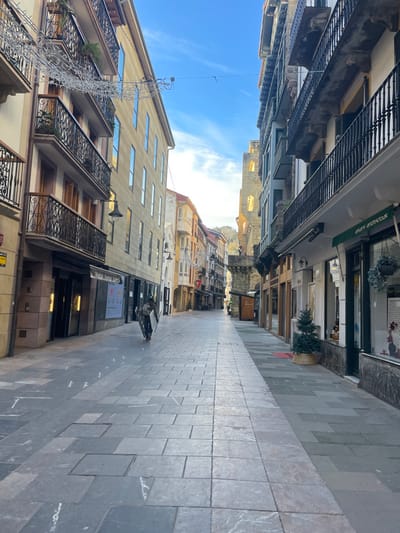 A narrow, pedestrianized street in Zarautz, Spain, is depicted under a clear blue sky. The street is lined on both sides by multi-story buildings featuring balconies with black wrought-iron railings and various window styles. Buildings on the left exhibit light-colored facades, some with reflective windows, and ground-floor businesses with dark storefronts. Christmas string lights are strung horizontally across the street, connecting buildings at an upper level. On the right, buildings feature darker facades; one ground-floor establishment displays a green awning with "arkami andreak" text and has a small Christmas tree in a wicker basket outside. Further down the street on the right, a prominent, tall stone tower or ruin with illuminated openings is visible. The street pavement consists of rectangular grey stone slabs of varying shades. One individual, wearing dark clothing and a backpack, walks away from the camera on the left side of the street, carrying a large, rectangular, light-colored panel vertically. Distant hills are visible at the far end of the street. Shadows fall across the left side of the pavement, indicating the direction of sunlight.