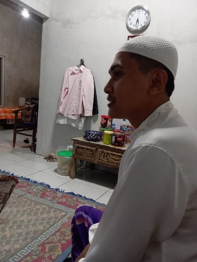 This is an interior scene located in Bulukandang, Indonesia. An adult male is seated in the right foreground, depicted in left profile. He wears a white knitted skullcap and a white long-sleeved shirt, with a purple sarong visible below. His mouth is slightly ajar. The floor is covered by a patterned rug on white tiles. In the midground, a carved wooden table holds multiple containers (red, yellow, dark blue) and a plastic water bottle. A light pink collared shirt and a dark garment hang on a wall hook behind the table. A round wall clock displaying approximately 12:35 is mounted on the upper wall. The background shows an open doorway to an adjacent room containing a chair and a table with an orange patterned cloth, illuminated by an exposed bulb. A green bucket is on the tiled floor near the wooden table. The walls are light-colored with visible staining.