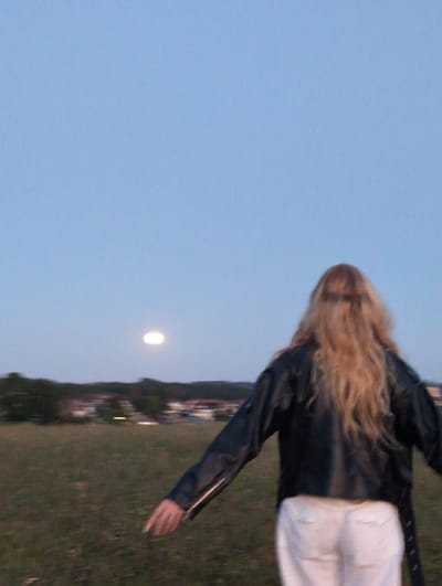 The image, taken in St. Peter, Germany, shows the back of a person with long, blonde hair wearing a black leather jacket and white pants. The person is standing in a grassy field, facing away from the camera. In the distance, a blurry landscape with trees and buildings is visible. A glowing, oval-shaped object, possibly the moon or a bright light, is suspended in the pale blue sky above the horizon. The overall lighting suggests it is dusk or twilight.