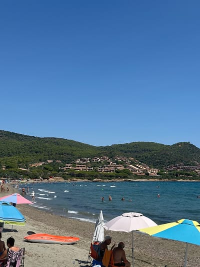 A wide shot captures a sun-drenched beach scene with a clear blue sky overhead and a calm blue sea. In the foreground, a sandy beach is dotted with beach umbrellas, chairs, and a bright orange kayak resting on the sand. Several people are visible on the beach, some sitting under umbrellas and others walking towards the water. The sea is populated with swimmers and a few specks of people further out. In the background, a lush green hillside is covered in dense trees and dotted with buildings, creating a picturesque backdrop. The overall impression is one of a peaceful and vibrant beach day in Villaputzu, Italy.