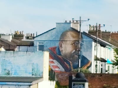 This image shows a mural of a man's face painted on the side of a building in Wallasey, United Kingdom. The mural covers a significant portion of the building's facade and depicts a man with dark skin, wearing a red and blue striped garment. The background is a clear blue sky with some rooftops and chimneys visible. In the foreground, parts of other buildings and street furniture, including a sign that reads "CUSTOMER PARKING ONLY," are present. The style of the mural is realistic, with attention to detail in the rendering of the man's features.