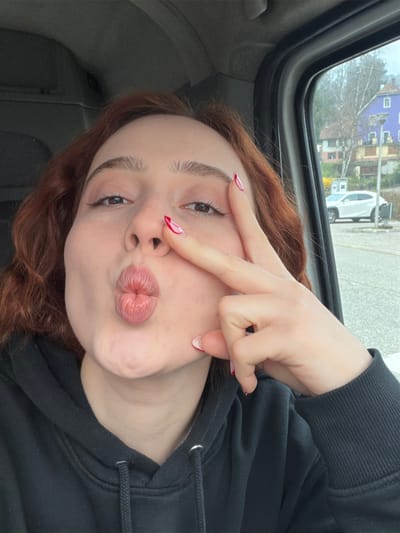 A selfie captures a young female subject inside a vehicle in Plaine, France. The subject, with reddish-brown wavy hair, wears a black hooded sweatshirt. Her lips are pursed in a "kissing" gesture, and her left hand (from the viewer's perspective) forms a "peace sign" with fingers extended and red and white manicured nails, positioned near her nose and cheek.
Through the vehicle's window on the right, an outdoor urban scene under an overcast daylight sky is visible. The environment includes a paved area, a parked white SUV, and multiple buildings. A prominent multi-story building, distinctively purple with a lighter ground floor, is visible on the right side of the background. Bare deciduous trees and distant hills are also present in the landscape, indicative of a colder season.