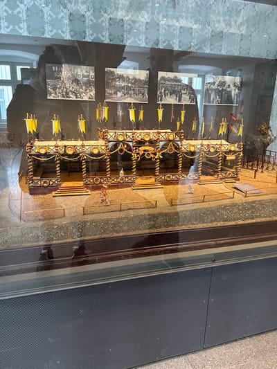 The image shows a diorama of a historic scene, displayed behind glass, likely within a museum or historical institution. The diorama is situated in Biberach an der Riß, Germany.

The diorama depicts an event featuring a crowd of people in front of a grandstand structure adorned with flags. Mounted on the glass display case, adjacent to the diorama, are black and white photographs depicting what appears to be the actual historical event that the diorama represents.

The setting is indoors, with a textured, decorative wall visible through the glass case. The floor nearby, to the side of the glass case, has a dark, perforated surface.