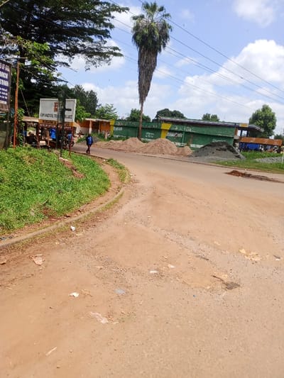 The image portrays a semi-urban roadside scene in Murang'a, Kenya, identified by a prominent white signboard on the left displaying "ADVENTIST TRA. COLLEGE MURANG'A CAMPUS." The foreground and mid-ground feature an unpaved, reddish-brown dirt road with visible ruts, potholes, and scattered plastic litter. A raised grassy bank with a concrete curb defines the left edge of the road. Overhead, numerous utility lines span a partly cloudy sky.

On the left, partially obscured by green foliage, a large white signboard and an older, rusted sign structure are visible. Beneath an adjacent shelter, several indistinct human figures are present, one appearing to be on a bicycle or motorcycle. A lone individual, dressed in a blue jacket and dark trousers, walks from left to right across the dirt road in the mid-ground.

To the right of the road, three distinct piles of construction materials are present: two lighter-colored piles of sand and one darker pile of gravel or crushed stone. A very tall, slender palm tree with a thick skirt of dead fronds dominates the right side of the image, extending into the sky. In the background, behind the construction materials, a long, low building painted green with a yellow stripe is visible. Further to the right in the background, additional structures and a partially obscured blue vehicle are present.